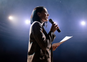 Mulher no palco segurando microfone e apresentado para plateia sua oratória.