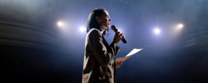 Mulher no palco segurando microfone e apresentado para plateia sua oratória.
