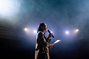 Mulher no palco segurando microfone e apresentado para plateia sua oratória.