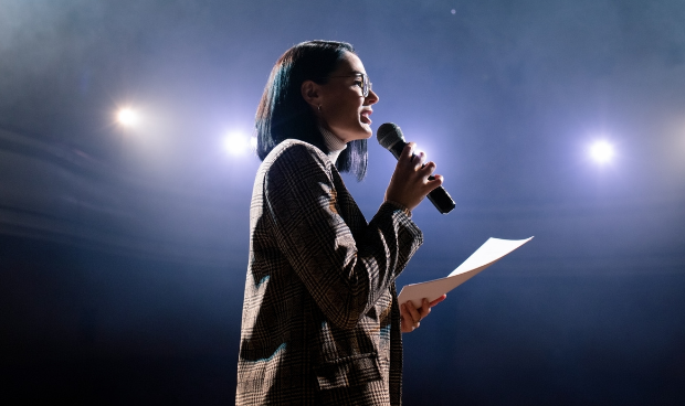 Mulher no palco segurando microfone e apresentado para plateia sua oratória.